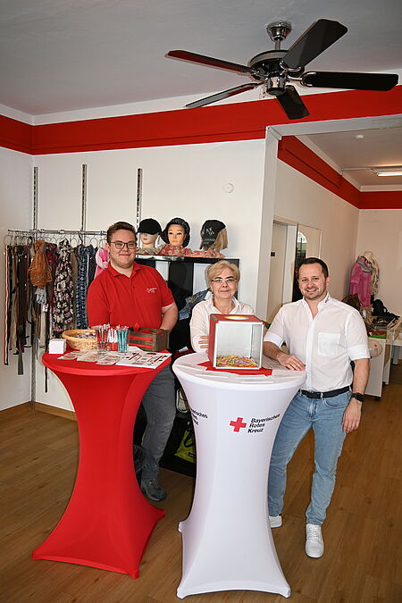 zwei Männer und eine Frau (Mitarbeiter des BRK) stehen zur Begrüßung der Kunden hinter Stehtischen